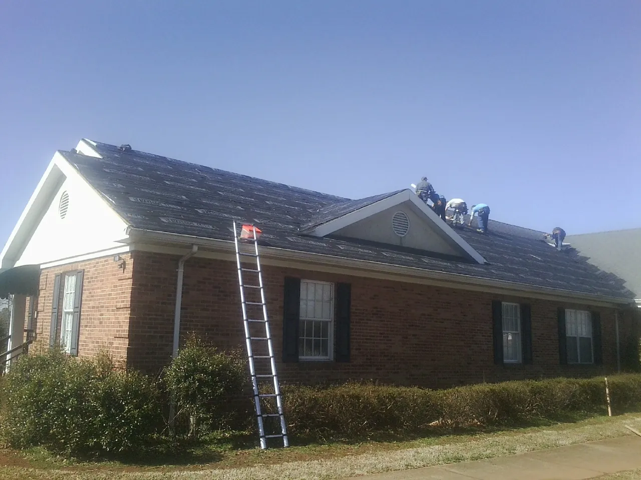 Crew tearing shingles off a brick ranch roof with a ladder set — Emergency Roof Repair work by our crew in Bokchito