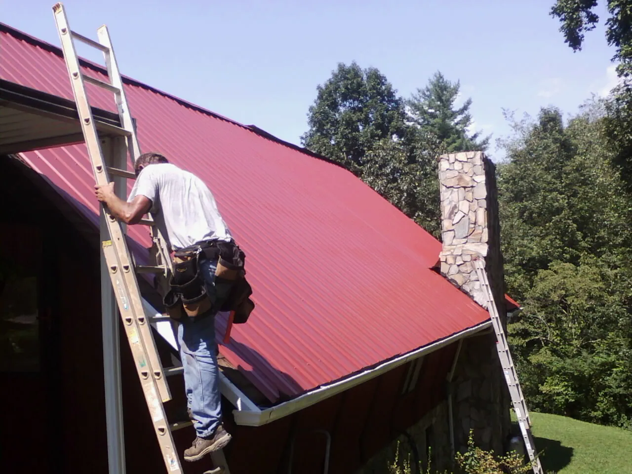 Roofer on a ladder beside a red metal roof and stone chimney — Soffit & Fascia Repair by Roofing Brothers in Bokchito